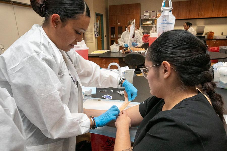 A student in lab coat demonstrates applying a bandage wrap on another student